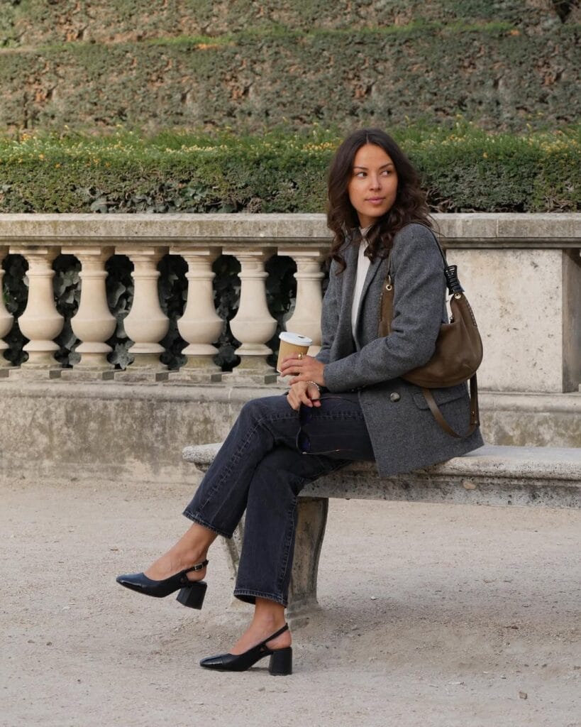 Woman in a grey blazer and black jeans, holding coffee on a stone bench; chic fall business casual outfit.