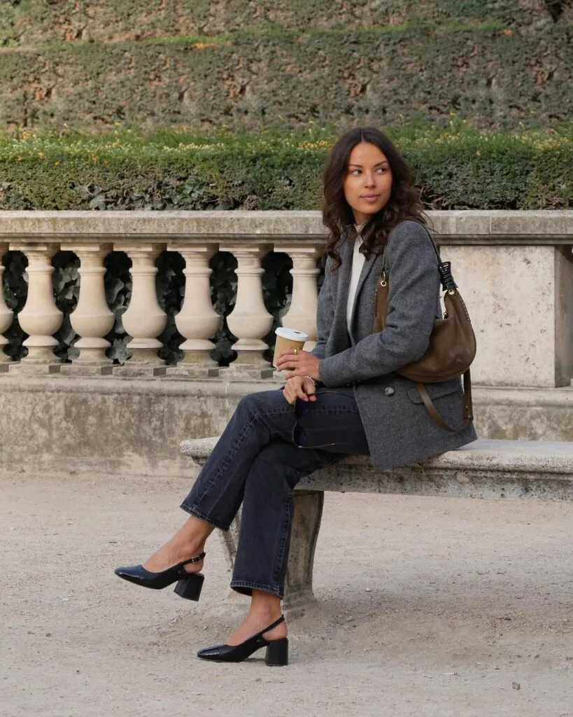 Woman in a grey blazer and black jeans, holding coffee on a stone bench; chic fall business casual outfit.