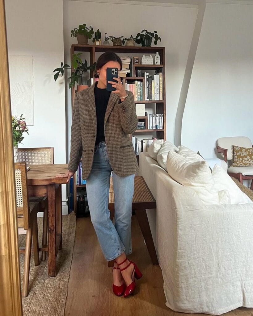 Woman in plaid blazer, black top, jeans, and red block-heel shoes, posing at home in a fall-ready outfit.