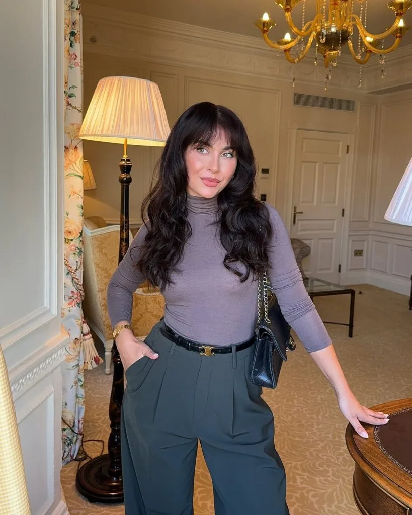 Woman in taupe turtleneck and pleated dark trousers with a black bag; fall business casual outfit in elegant room.
