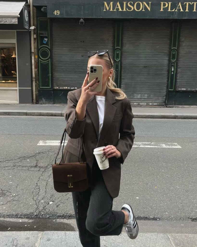 Woman in brown blazer, white tee, dark jeans, and sneakers, holding coffee; business casual style for fall.