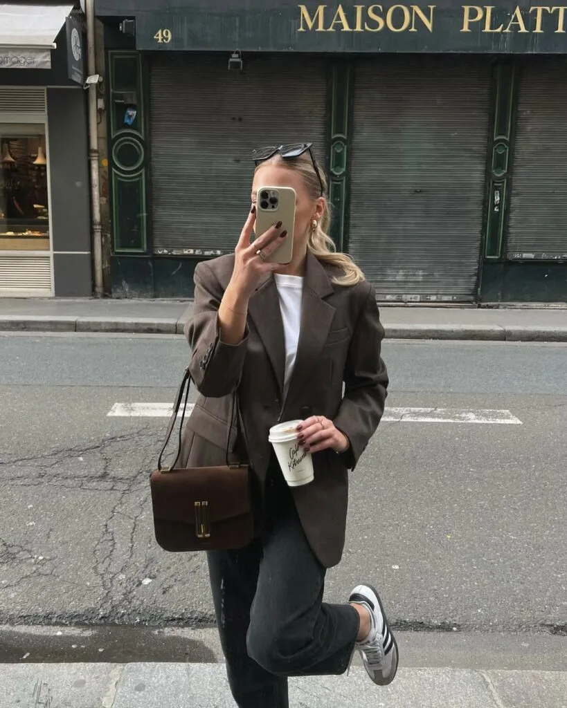 Woman in brown blazer, white tee, dark jeans, and sneakers, holding coffee; business casual style for fall.