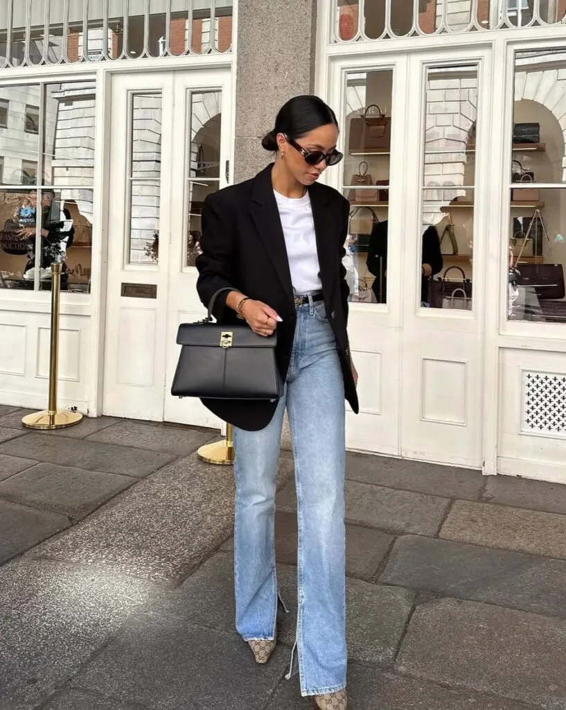Woman in black blazer, white top, light-wash jeans, and pointed heels; fall business casual outfit outside shop.
