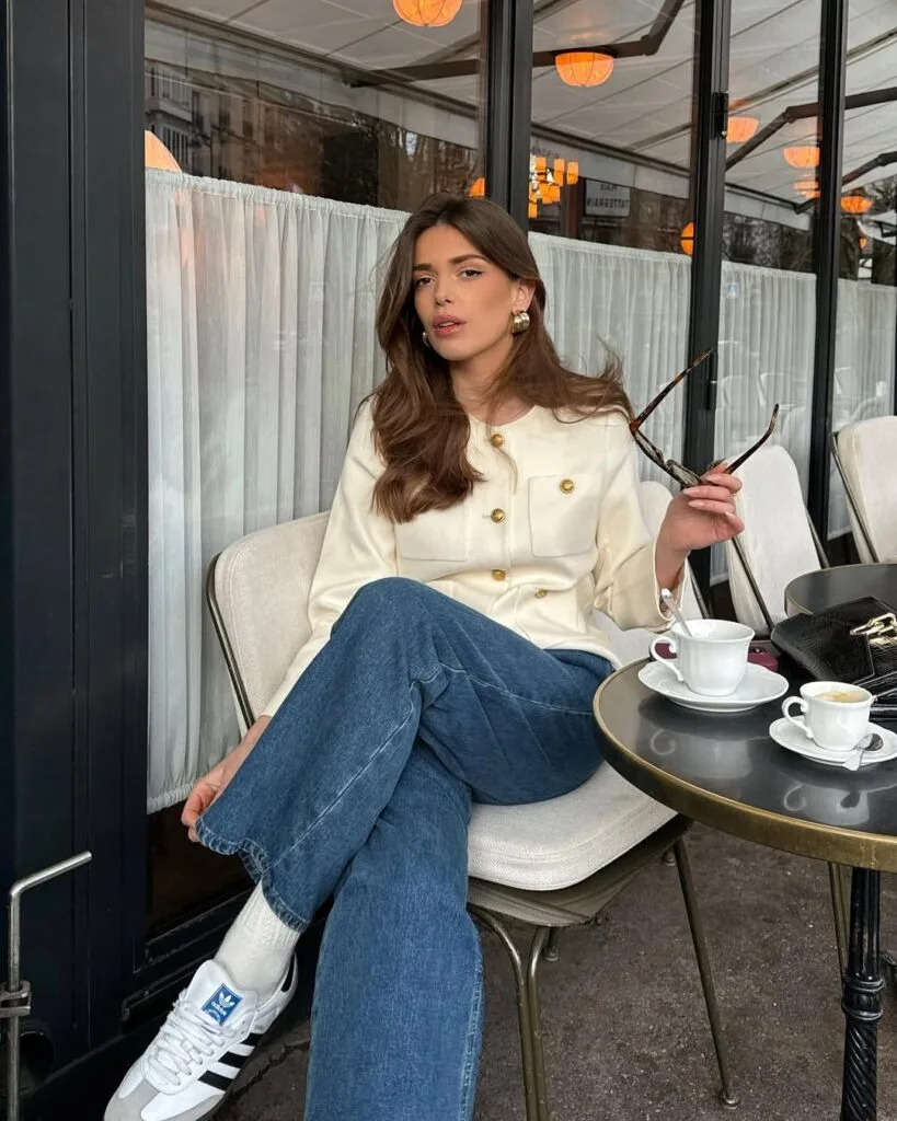  Woman in cream jacket, blue jeans, and Adidas sneakers, sitting at a Parisian cafe; business casual look.