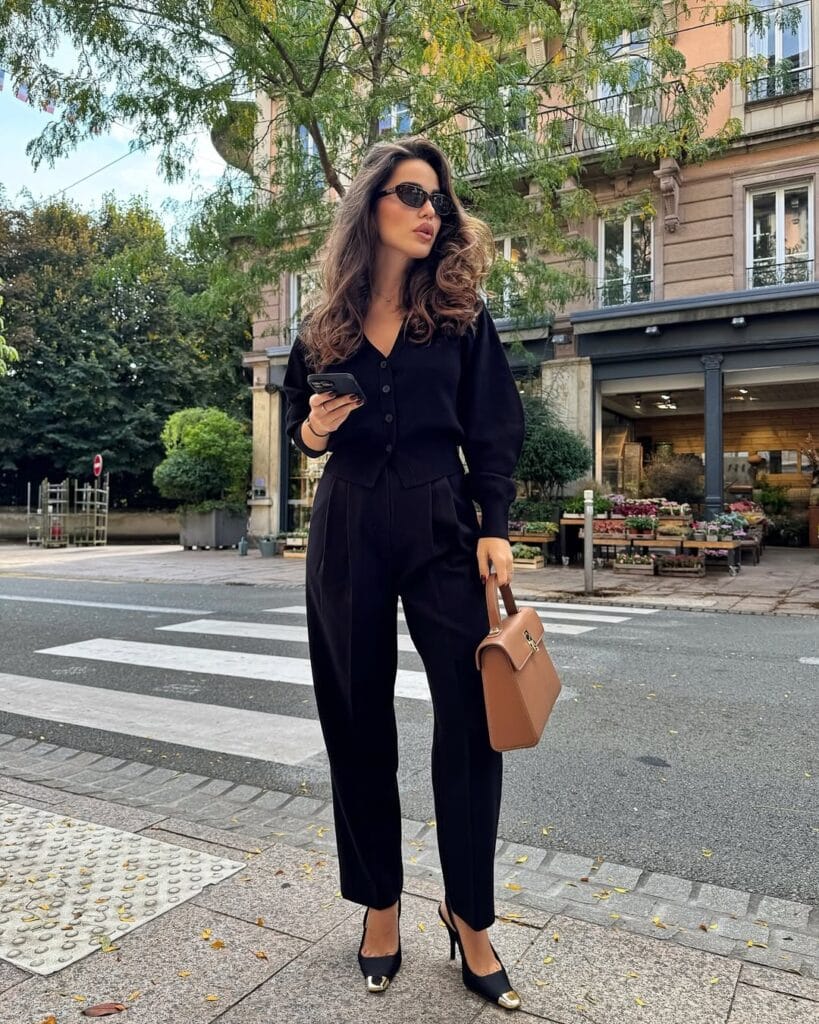 Woman in all-black cardigan and trousers, holding tan bag, crossing street; business casual fall outfit.
