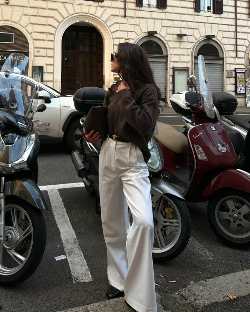 Woman in brown sweater and white wide-leg pants, standing near scooters; chic fall business casual outfit.
