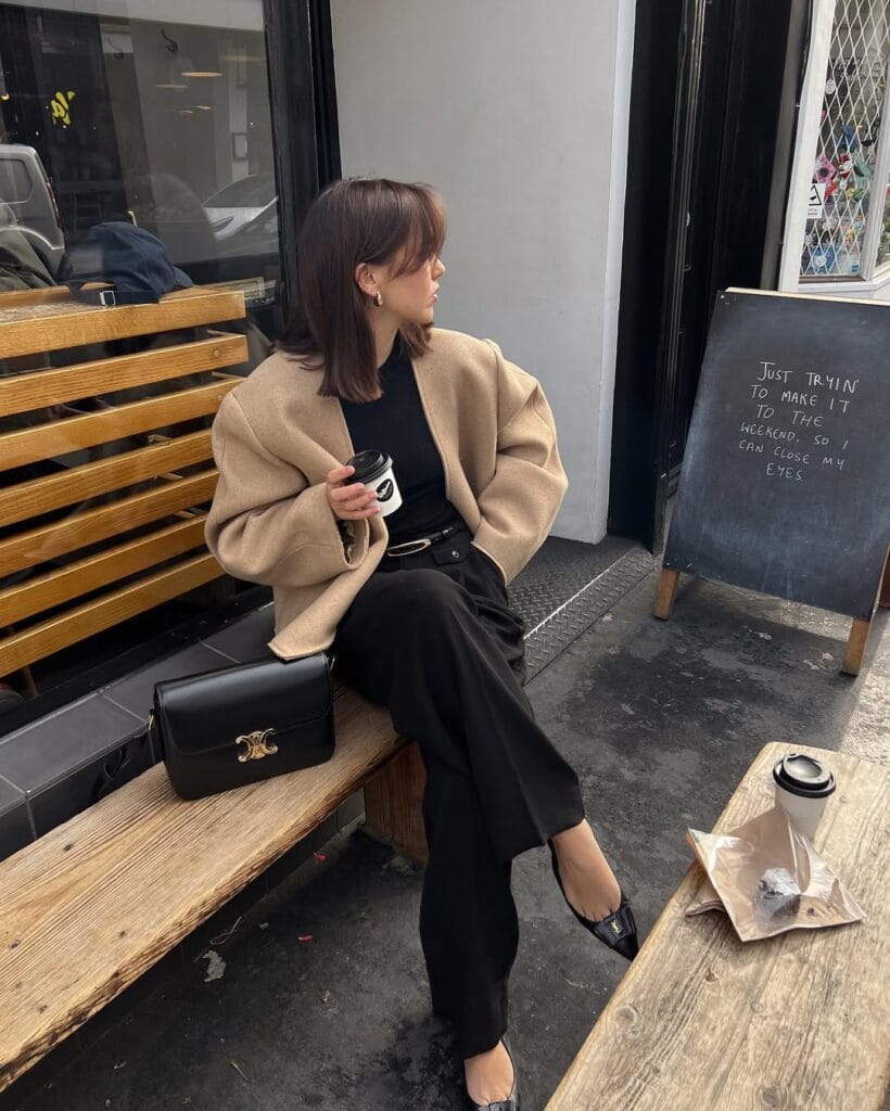 Woman in camel coat and all black outfit, holding coffee, sitting on bench; fall business casual look.
