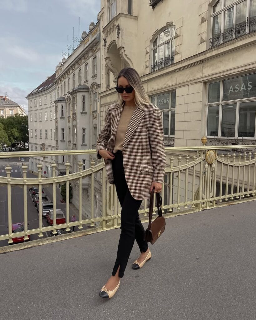 Woman in checked blazer, camel sweater, black pants, and cap-toe flats, walking on city bridge; fall business casual outfit.
