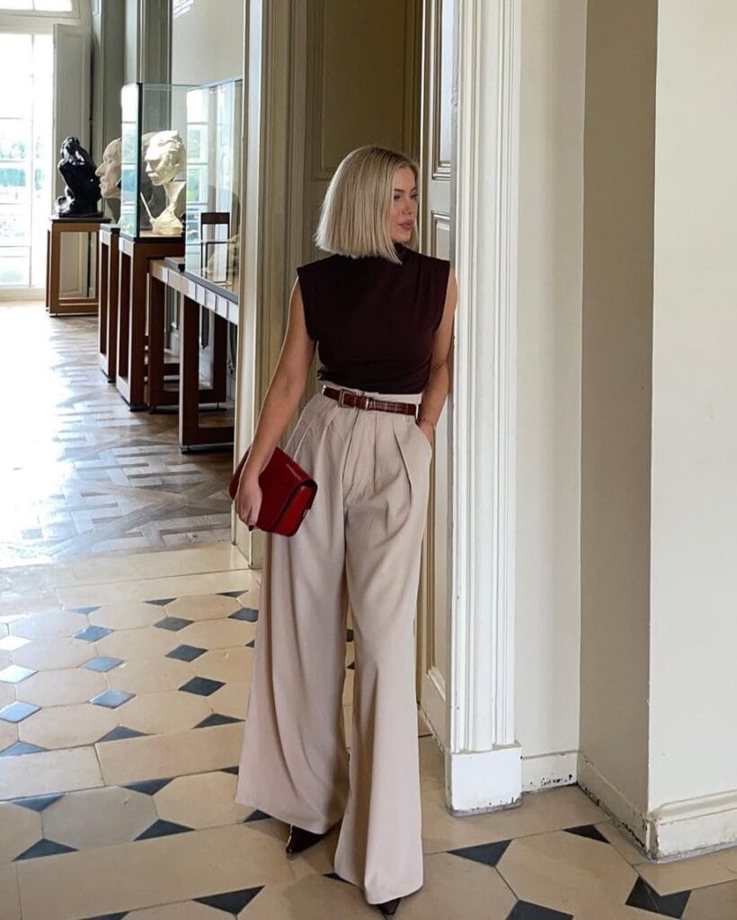 Woman in sleeveless brown top and beige wide-leg trousers, with red clutch, standing in an art gallery
