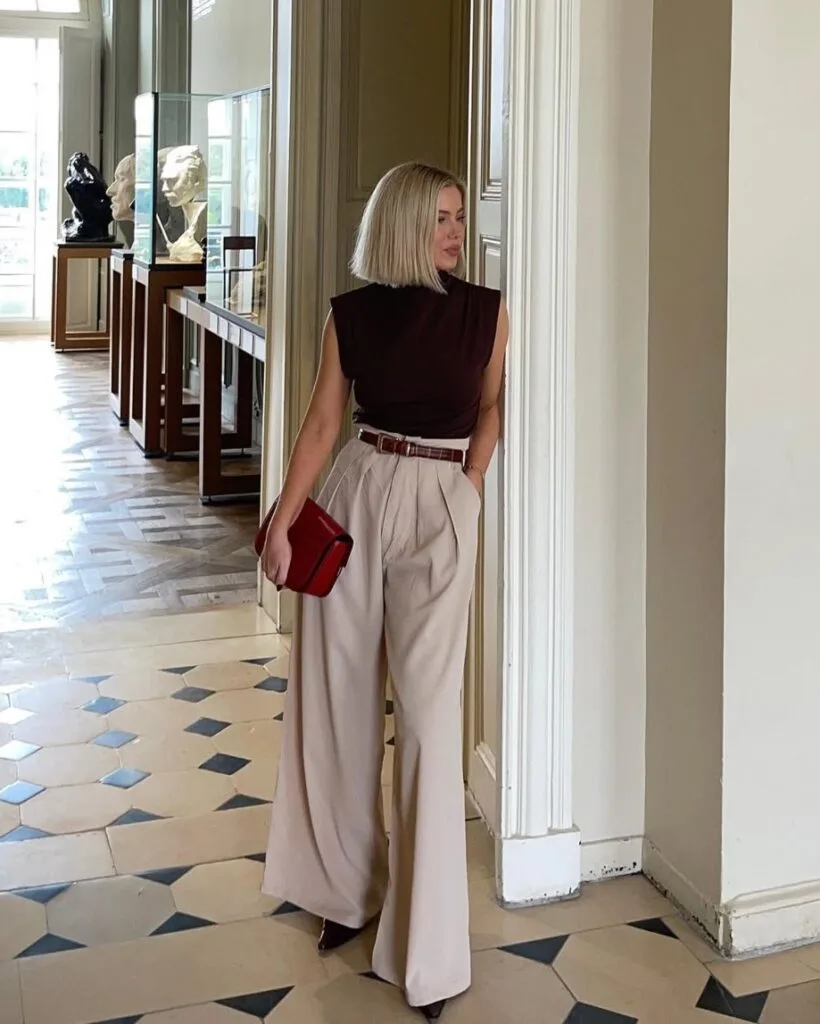 Woman in sleeveless brown top and beige wide-leg trousers, with red clutch, standing in an art gallery
