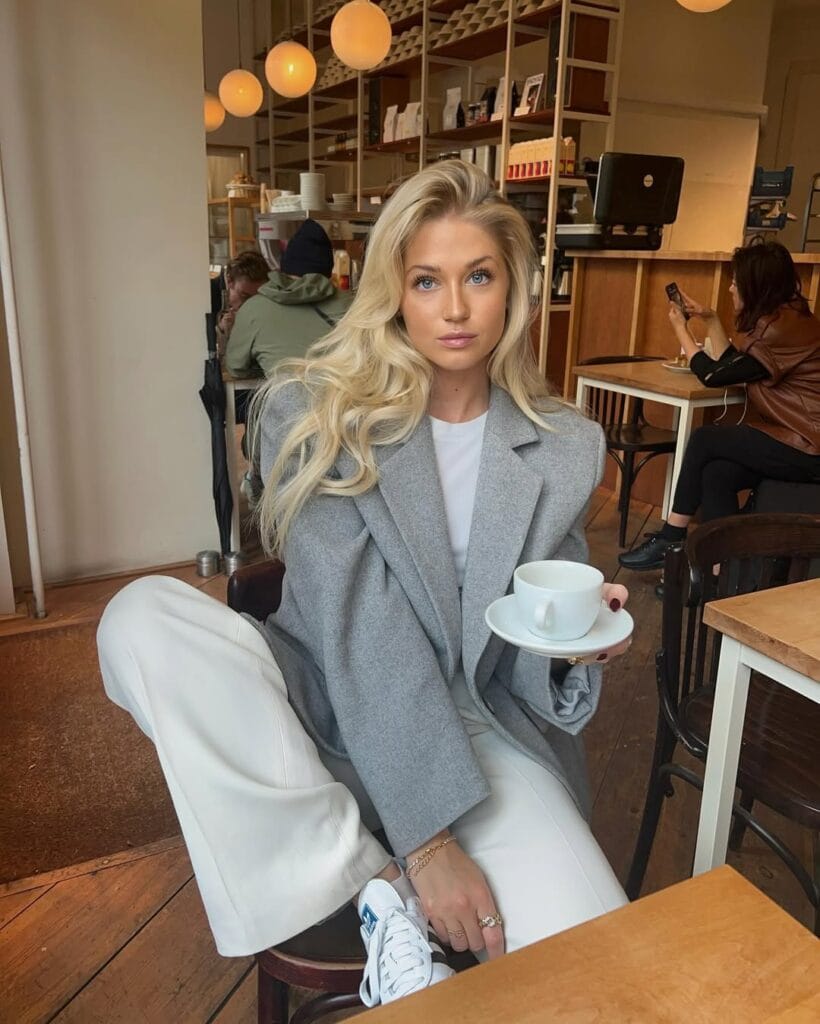 Woman in oversized grey blazer, white tee, and wide-leg pants with sneakers, sitting in a cozy cafe.
