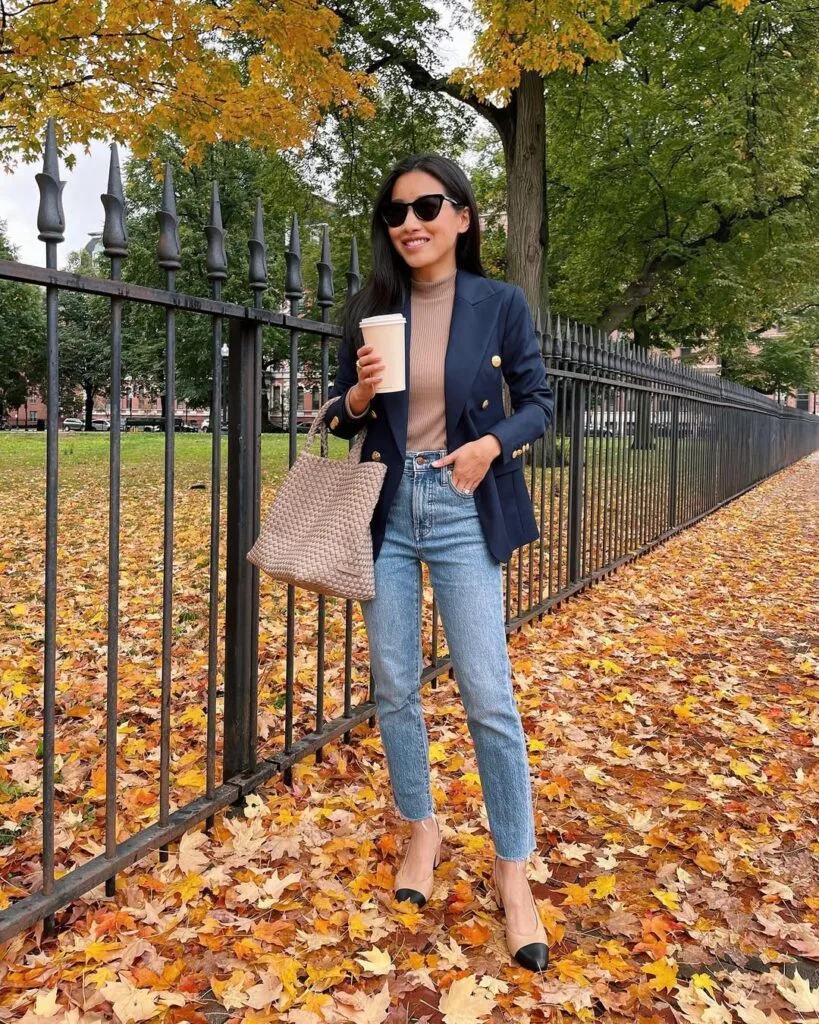 Woman in navy blazer, taupe turtleneck, and jeans, standing on autumn leaves, holding coffee, with a woven tote.