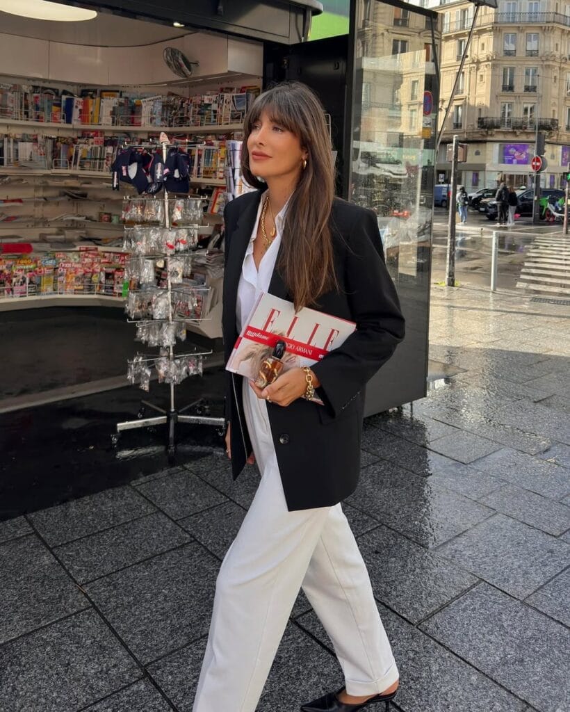 Woman in black blazer, white trousers, and white shirt, walking outdoors with a magazine and gold jewelry.
