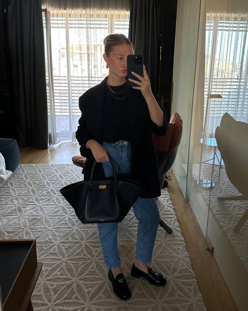 Woman in black blazer and top, blue jeans, black loafers, taking a mirror selfie with a structured black bag.
