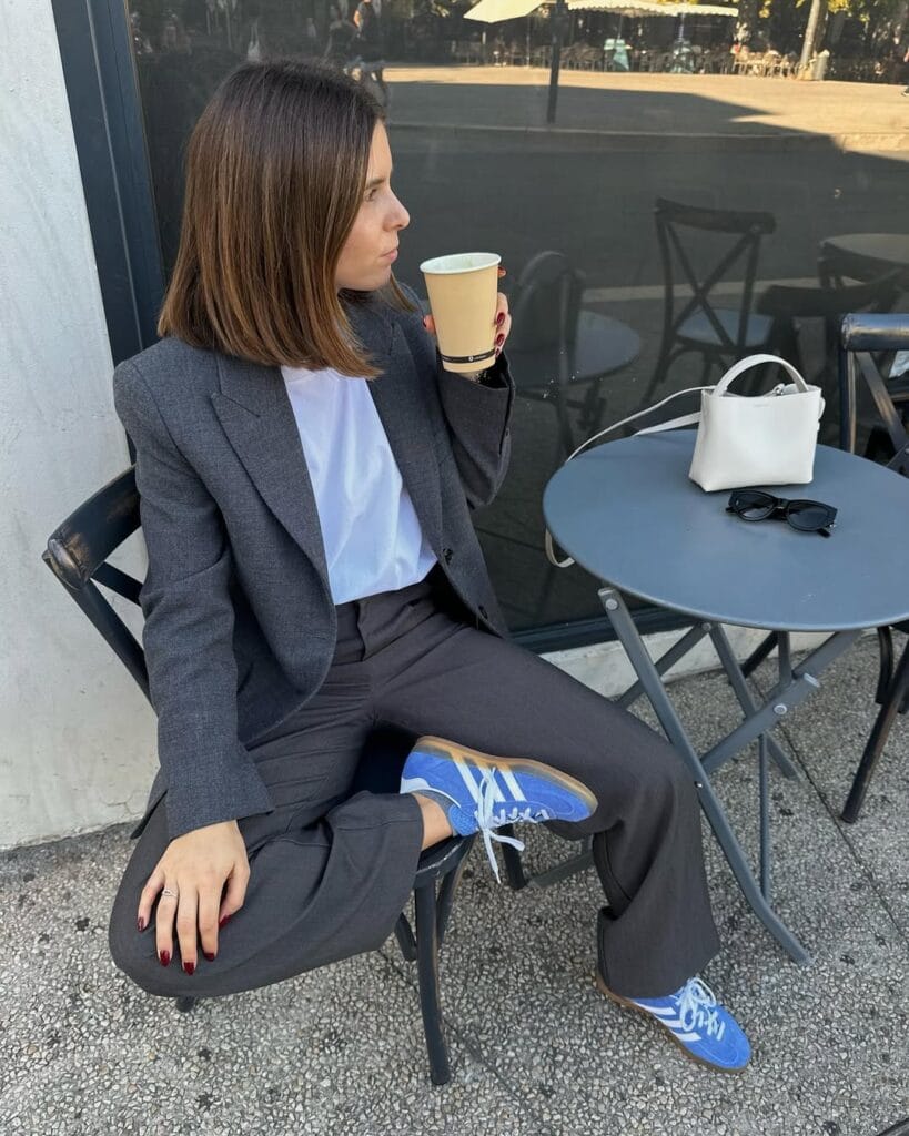 Woman in charcoal blazer and pants, white tee, blue sneakers, seated outdoors with coffee and white mini bag.
