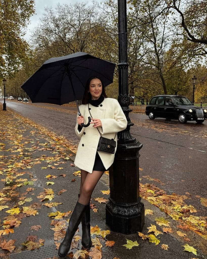 Woman in cream coat and black rain boots holding umbrella on a rainy street, classic rainy day outfit with fall leaves.
