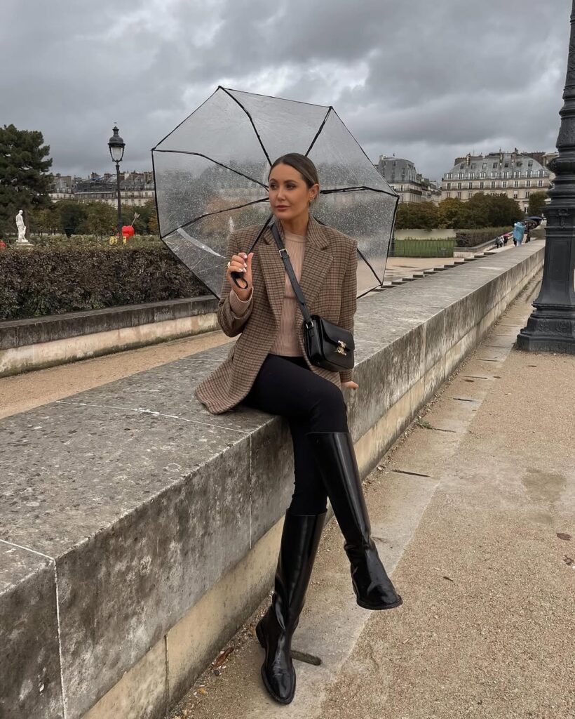 Woman in taupe checkered blazer, tan sweater, black pants, tall boots, clear umbrella; classic rainy day outfit for city stroll.
