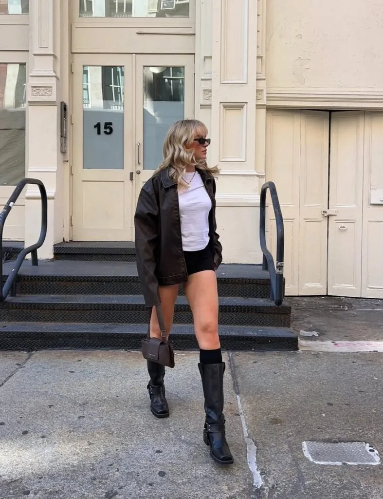 Woman in black leather jacket, white tee, micro shorts, knee boots, city setting; cool rainy day outfit idea.
