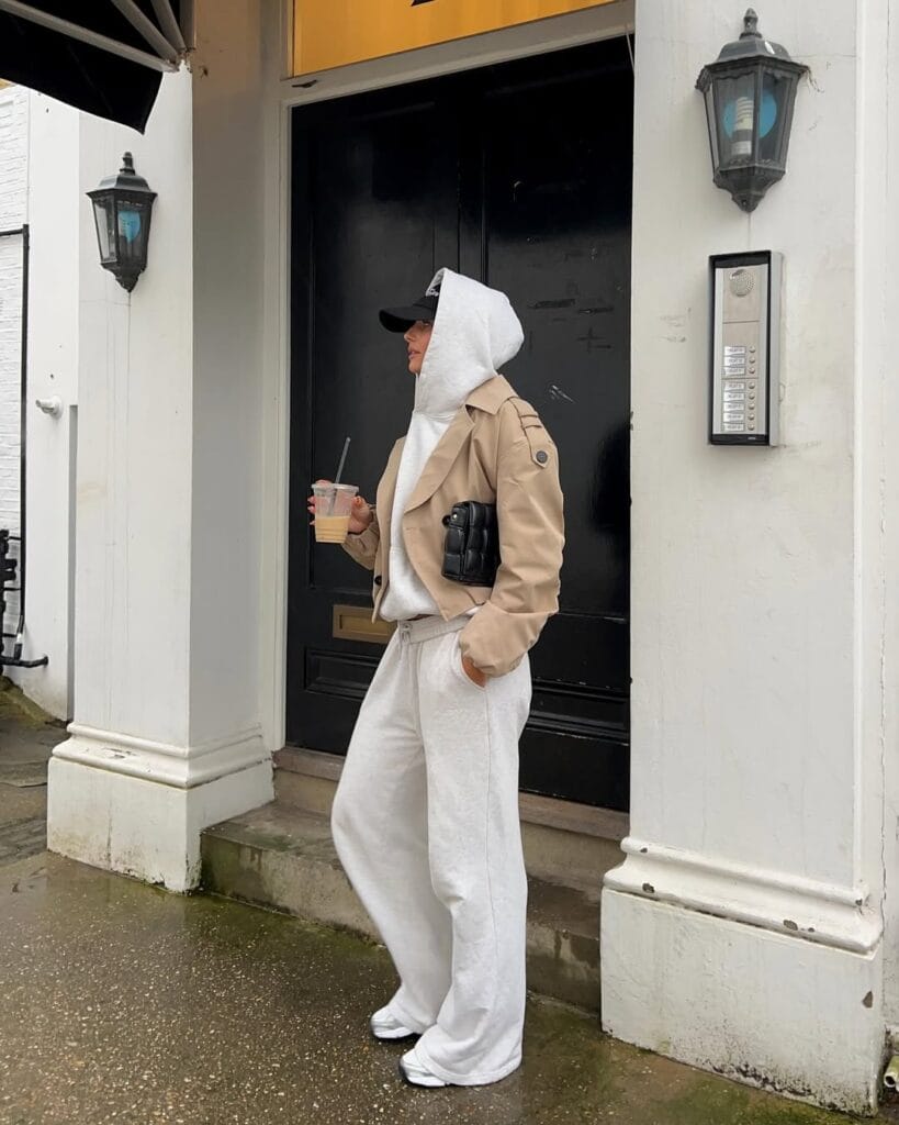 Woman in grey hoodie, beige cropped trench, white sweats, cap, holding coffee; sporty rainy day outfit.
