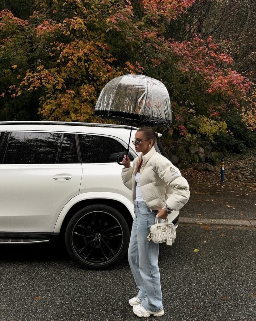 Woman in white puffer, tank, light jeans, sneakers, clear umbrella; chic rainy day outfit, autumn street.
