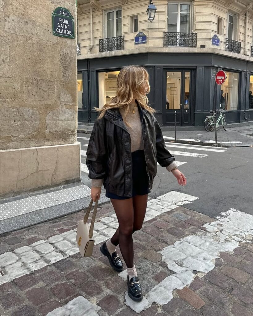 Woman in black bomber, tan sweater, black shorts, tights, loafers; rainy day outfit for city errands.

