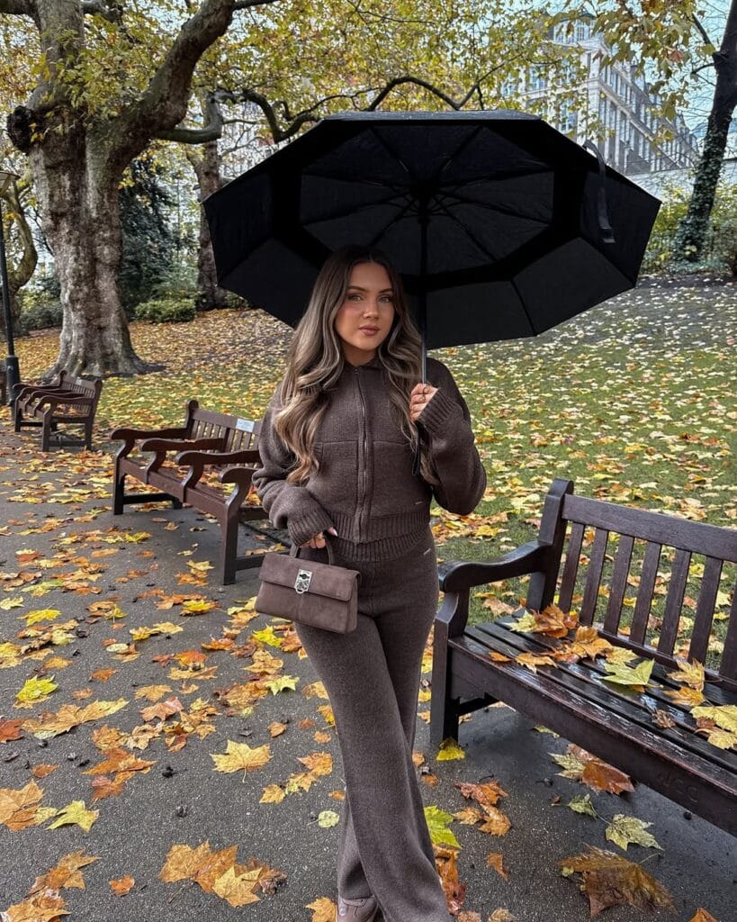 Woman in brown knit zip-up, matching pants, umbrella; cozy and chic rainy day outfit in leafy park.
