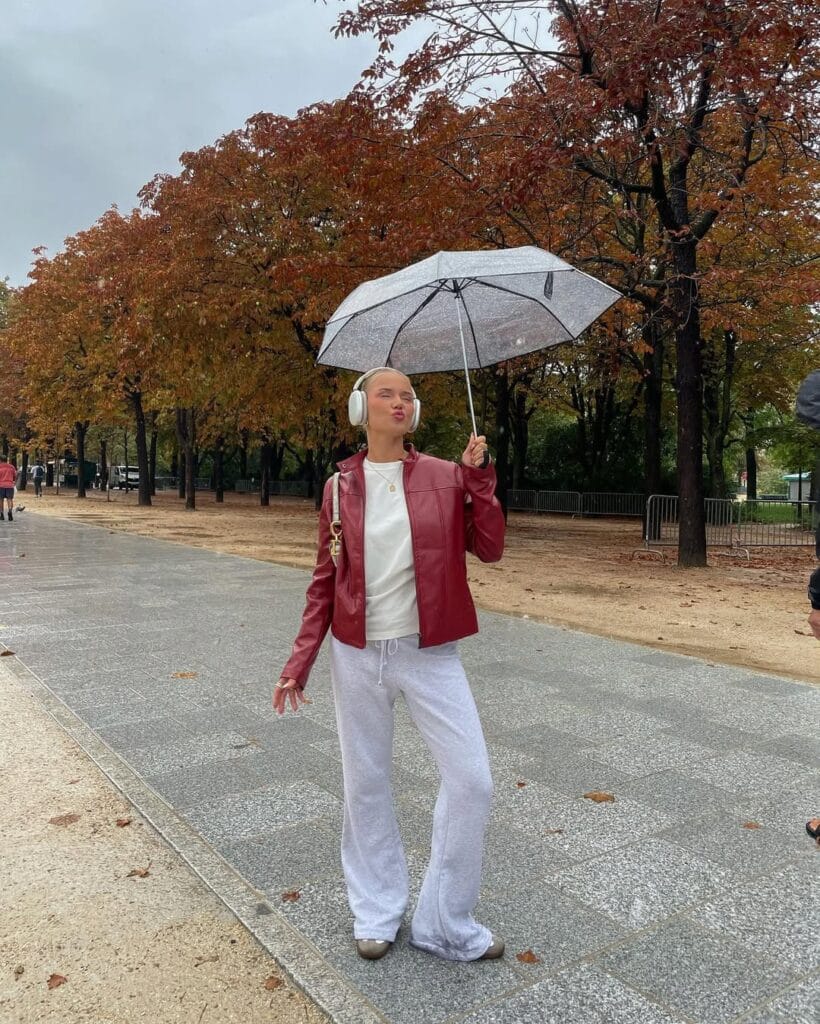 Woman in red jacket, white lounge pants, umbrella and earmuffs, playful rainy day outfit on city sidewalk.
