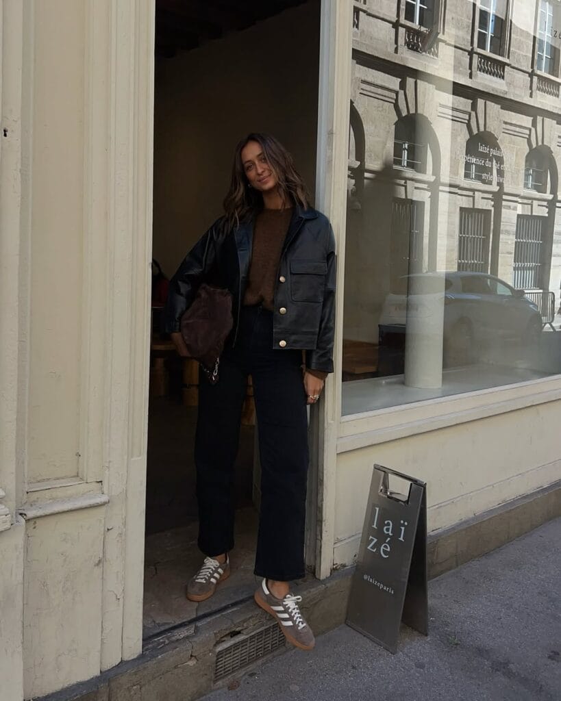 Woman in black cropped leather jacket, brown knit, straight jeans, sneakers; casual rainy day outfit.