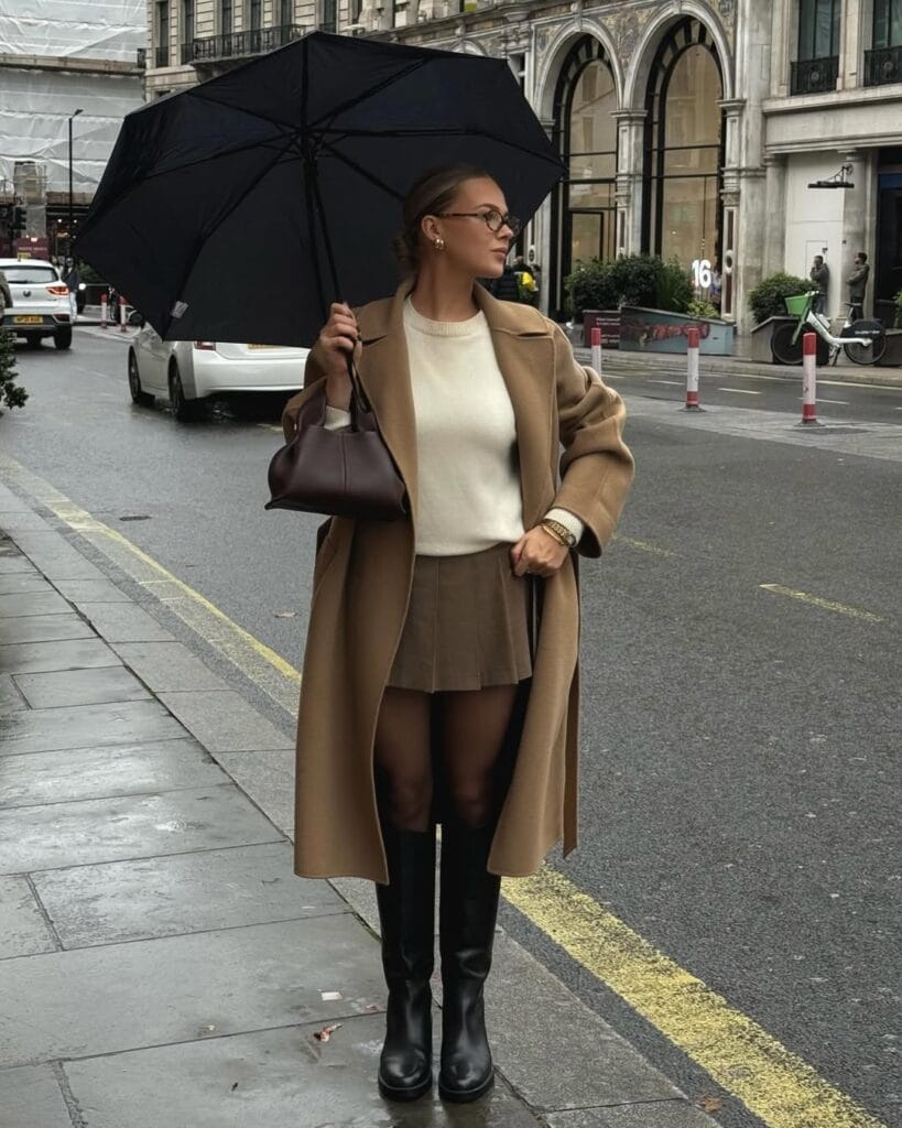 Woman in camel coat, cream sweater, pleated mini, knee boots, umbrella; classy rainy day outfit.
