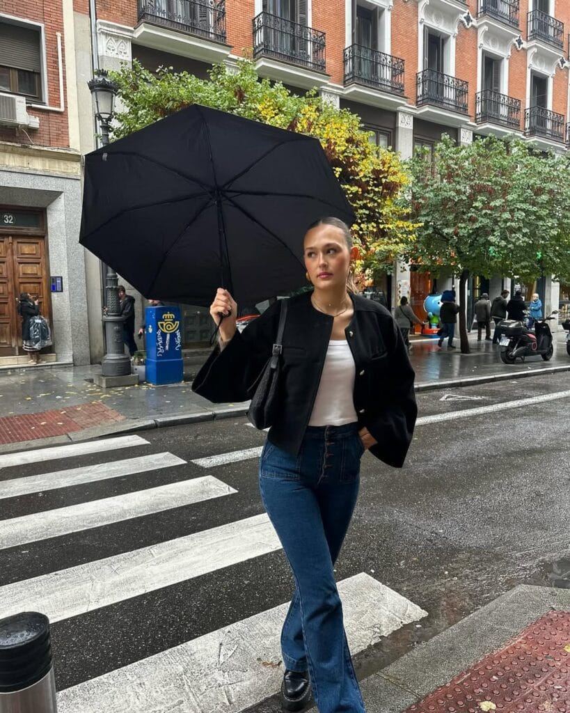 Woman in black cropped jacket, white top, bootcut jeans, umbrella; easy rainy day outfit for city walk.
