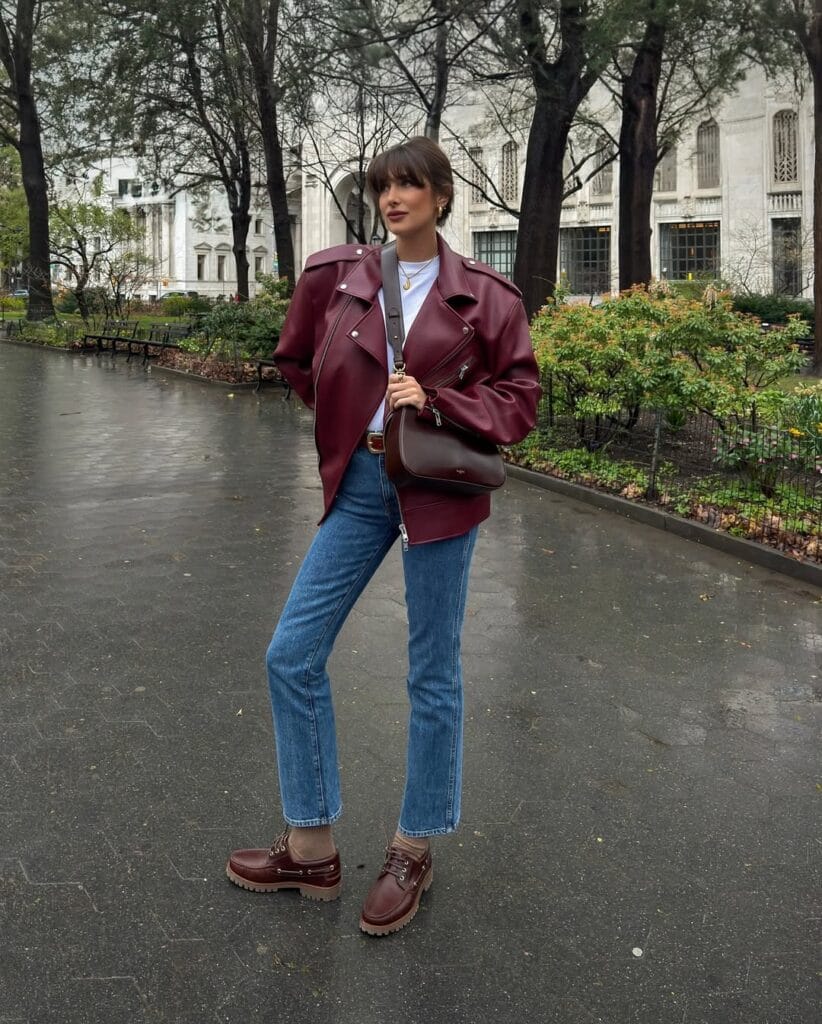Woman in burgundy leather moto jacket, blue jeans, brown boots; edgy rainy day outfit in park.
