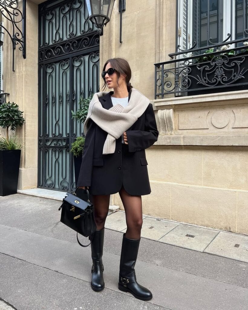Woman in black blazer, knit scarf and tall boots, stylish rainy day outfit for city walks.
