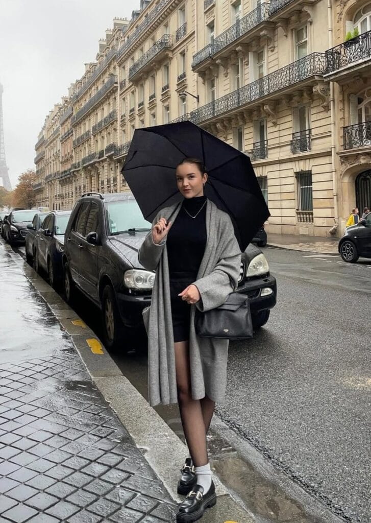Woman in grey long cardigan, black outfit, loafers and umbrella on wet Paris street, classic rainy day outfit.

