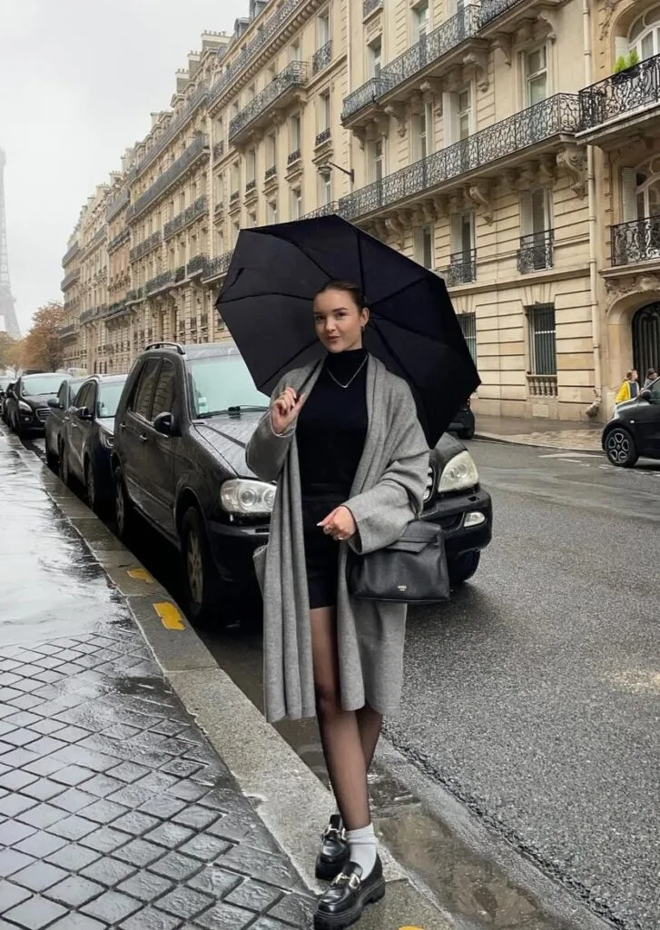 Woman in grey long cardigan, black outfit, loafers and umbrella on wet Paris street, classic rainy day outfit.
