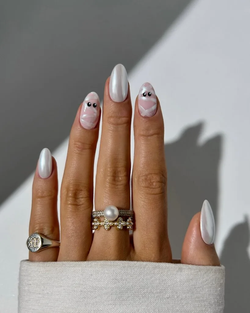 Pearl white Halloween nails with pink mummy wrap art and black eyes on almond nails.
