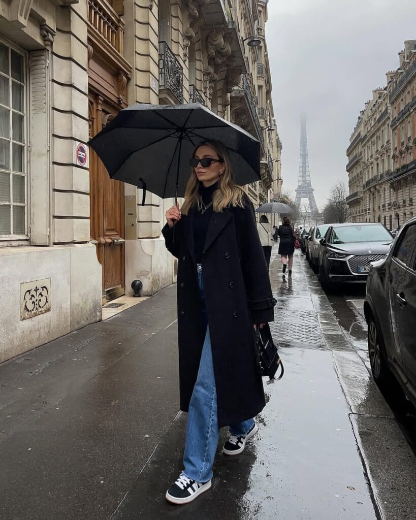 Woman in long black coat, blue wide-leg jeans, sneakers and umbrella, minimalist rainy day outfit.

