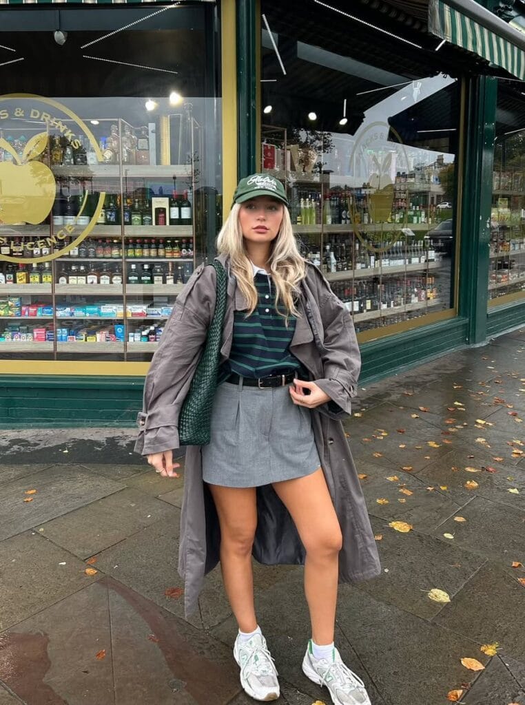 Woman in oversized trench, tennis skirt, green cap and sneakers, sporty rainy day outfit in city.
