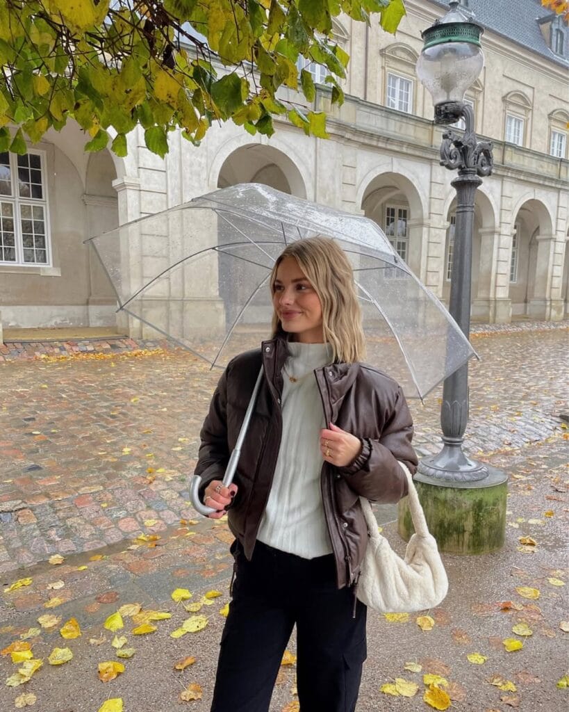 Woman in brown bomber jacket, white sweater, black pants and clear umbrella, chic rainy day outfit.