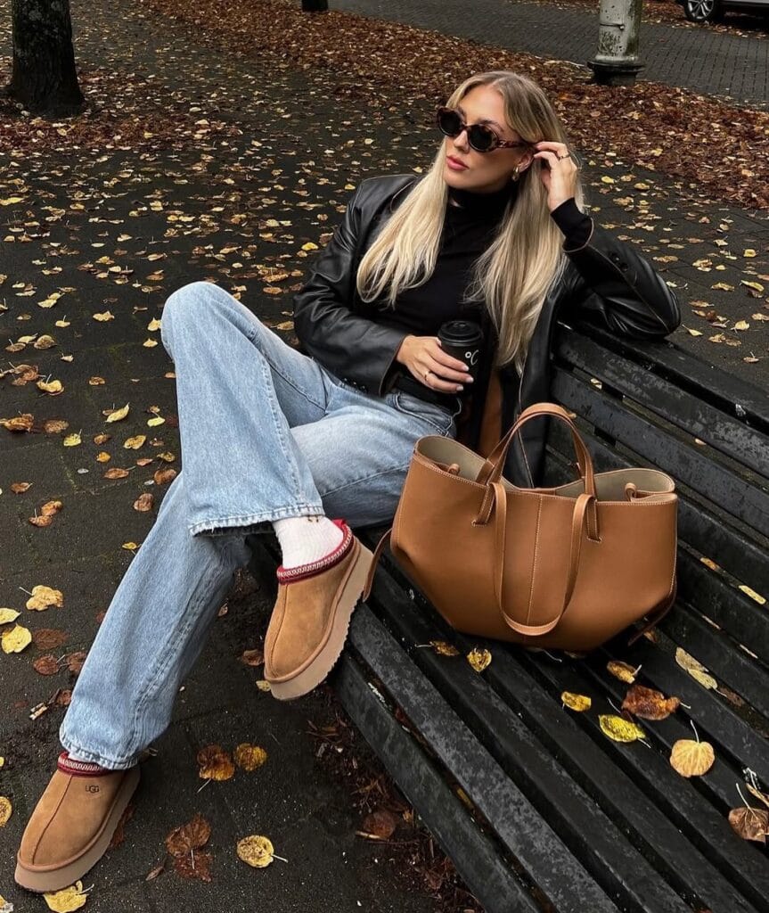 Woman in black leather jacket, jeans, tan UGGs, sunglasses on bench, relaxed rainy day outfit for fall.

