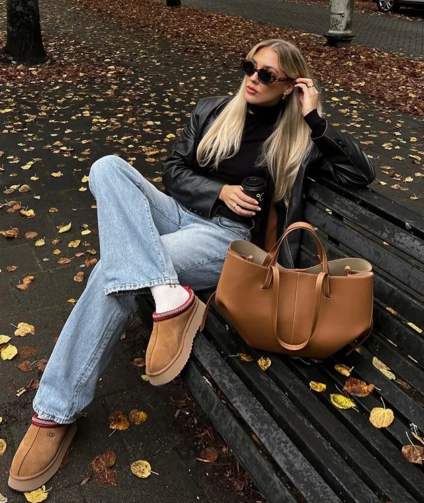 Woman in black leather jacket, jeans, tan UGGs, sunglasses on bench, relaxed rainy day outfit for fall.
