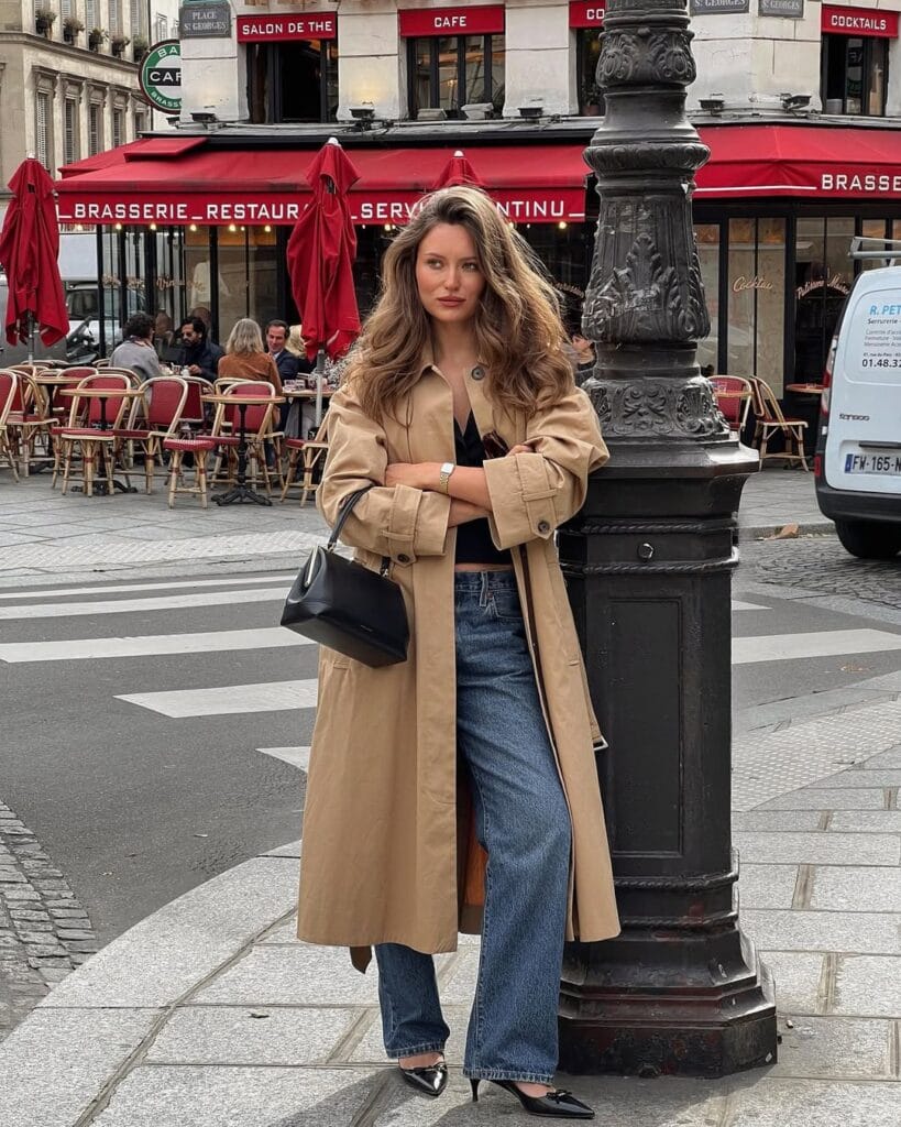 Woman in tan trench coat with blue jeans and black heels outside Parisian café; classic French style.