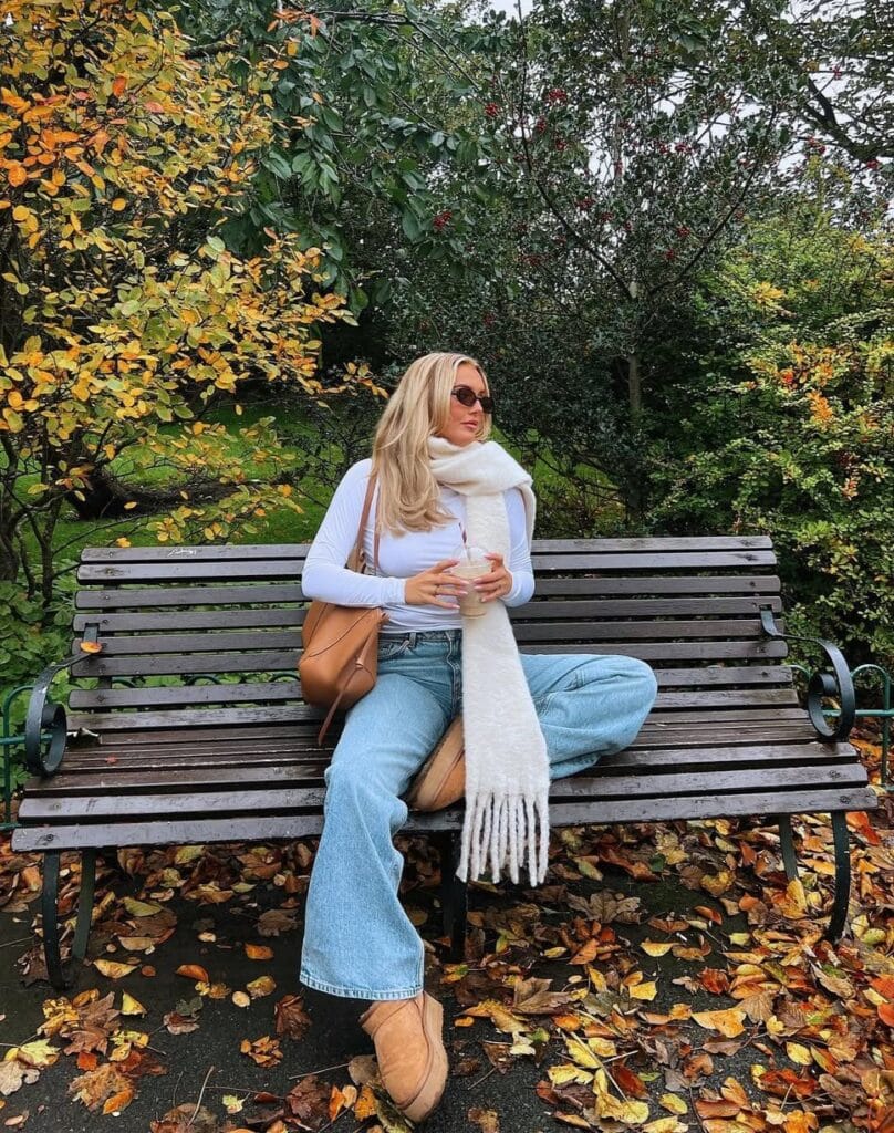 Woman on park bench in white top, scarf, jeans, and Ugg boots with fall leaves; cozy Ugg outfit.