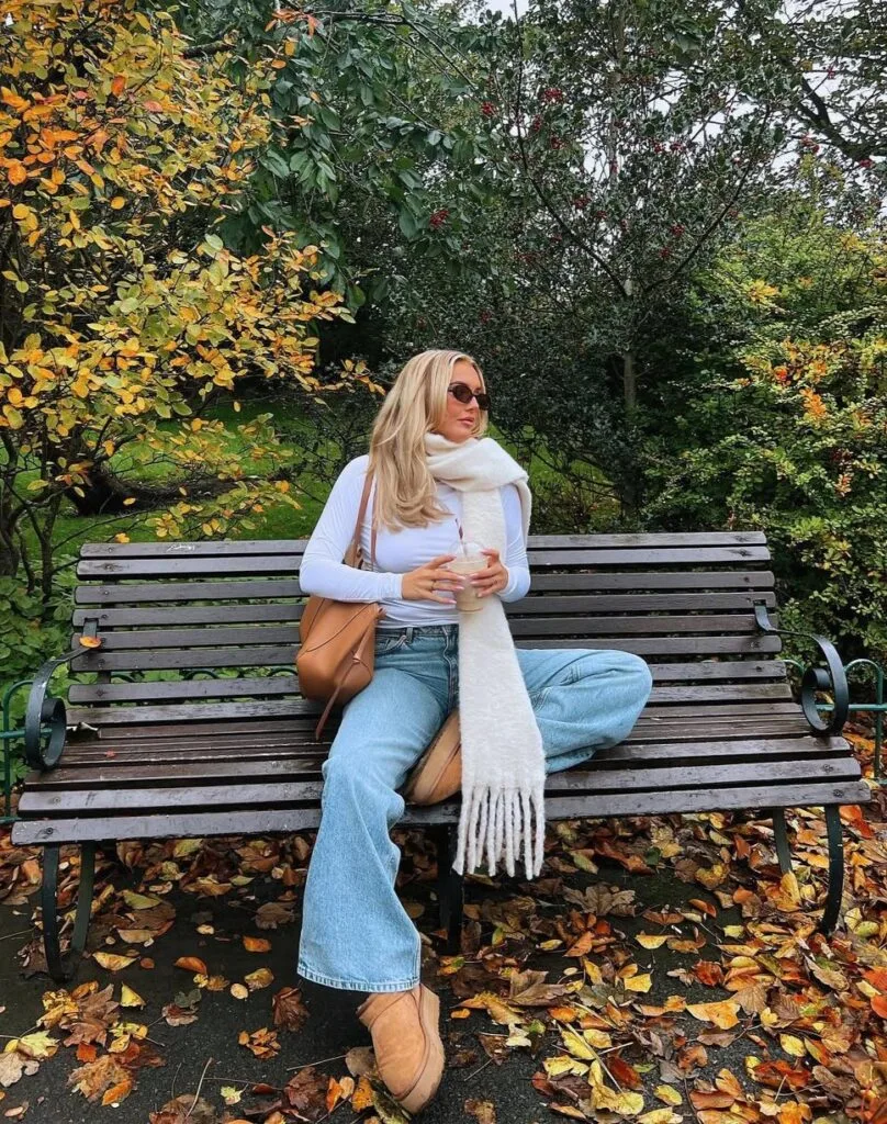 Woman on park bench in white top, scarf, jeans, and Ugg boots with fall leaves; cozy Ugg outfit.
