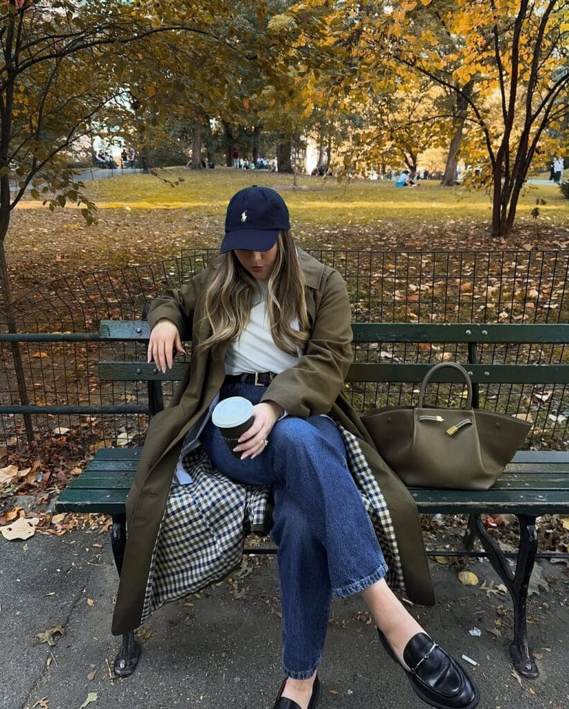 Woman in olive trench coat, jeans, loafers, and cap sitting on a park bench