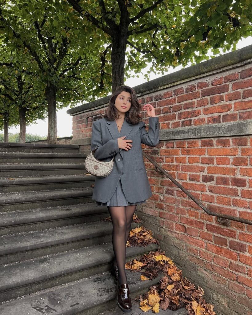 Woman in gray blazer, matching mini skirt, sheer tights, brown loafers; fall loafers outfit on stone steps.
