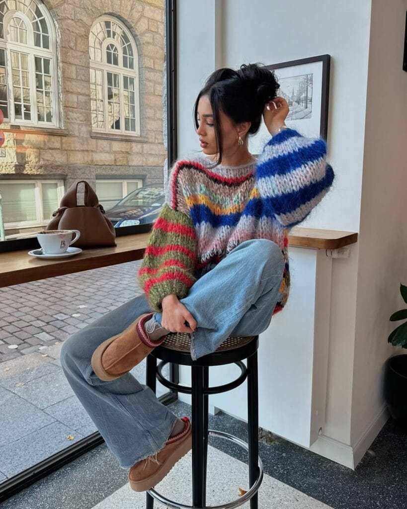 Woman in colorful striped sweater and Ugg boots, sitting on bar stool by café window; fun Ugg outfit idea.