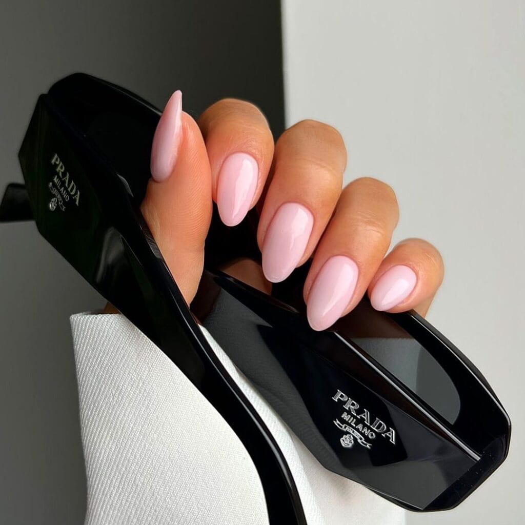 Alt text: Glossy baby pink almond neutral nails, holding black sunglasses, feminine and trendy.
