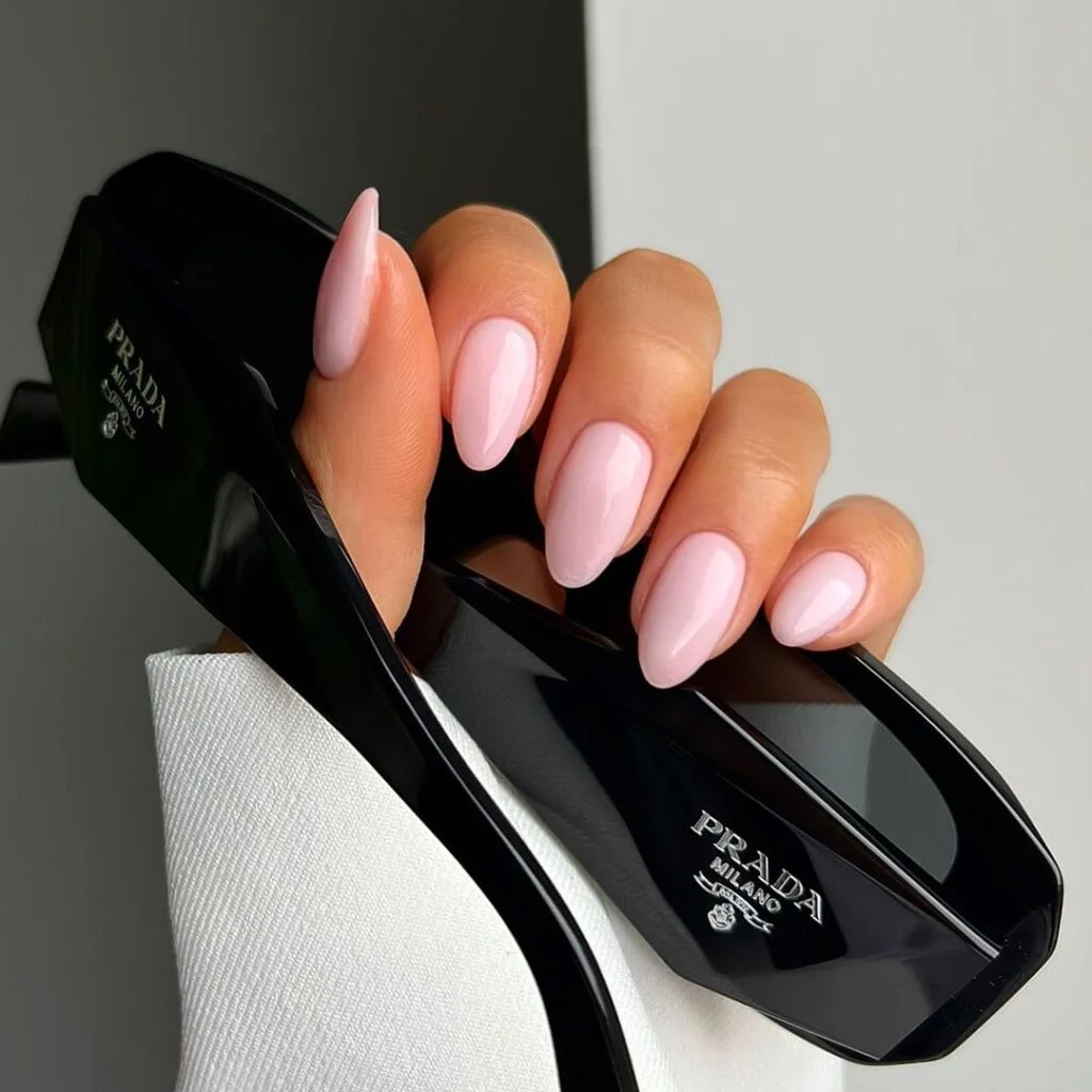 Alt text: Glossy baby pink almond neutral nails, holding black sunglasses, feminine and trendy.
