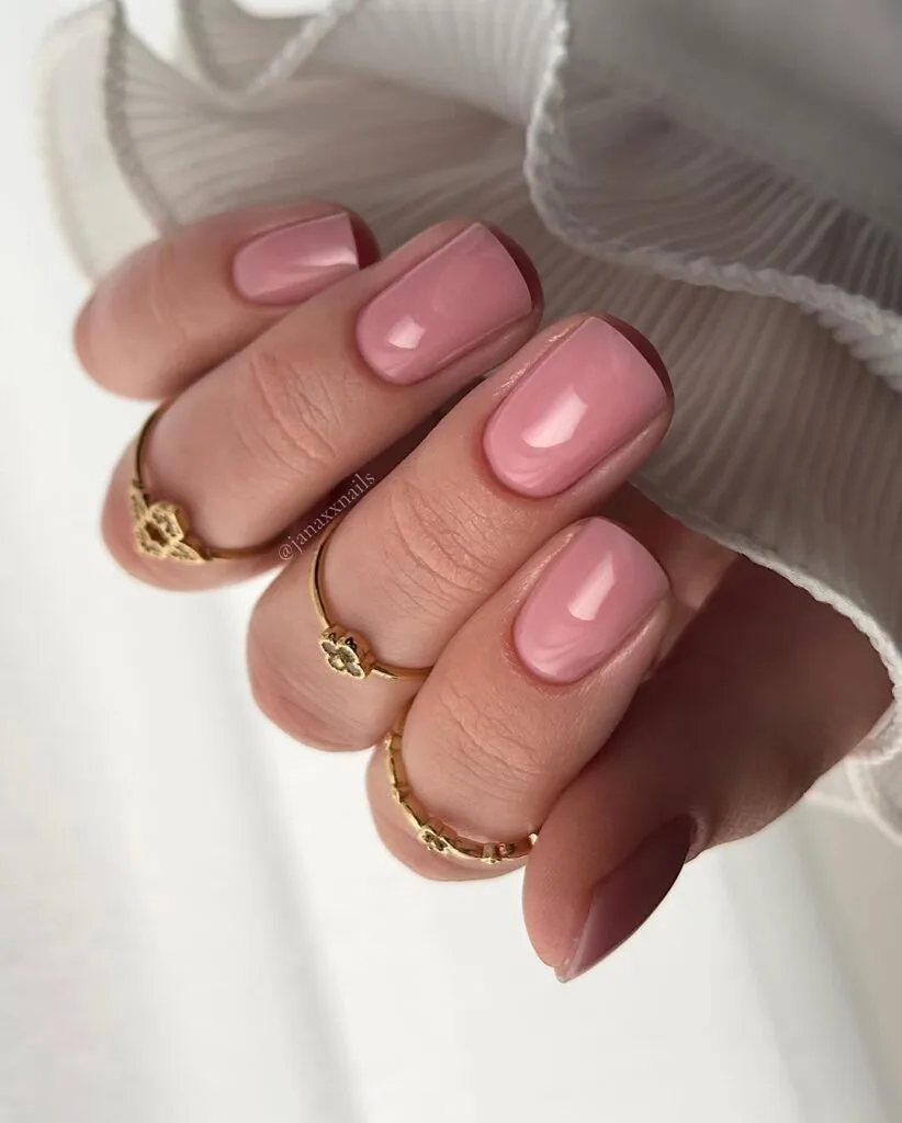 Alt text: Short dusty rose neutral nails, square shape, glossy finish, styled with gold rings.