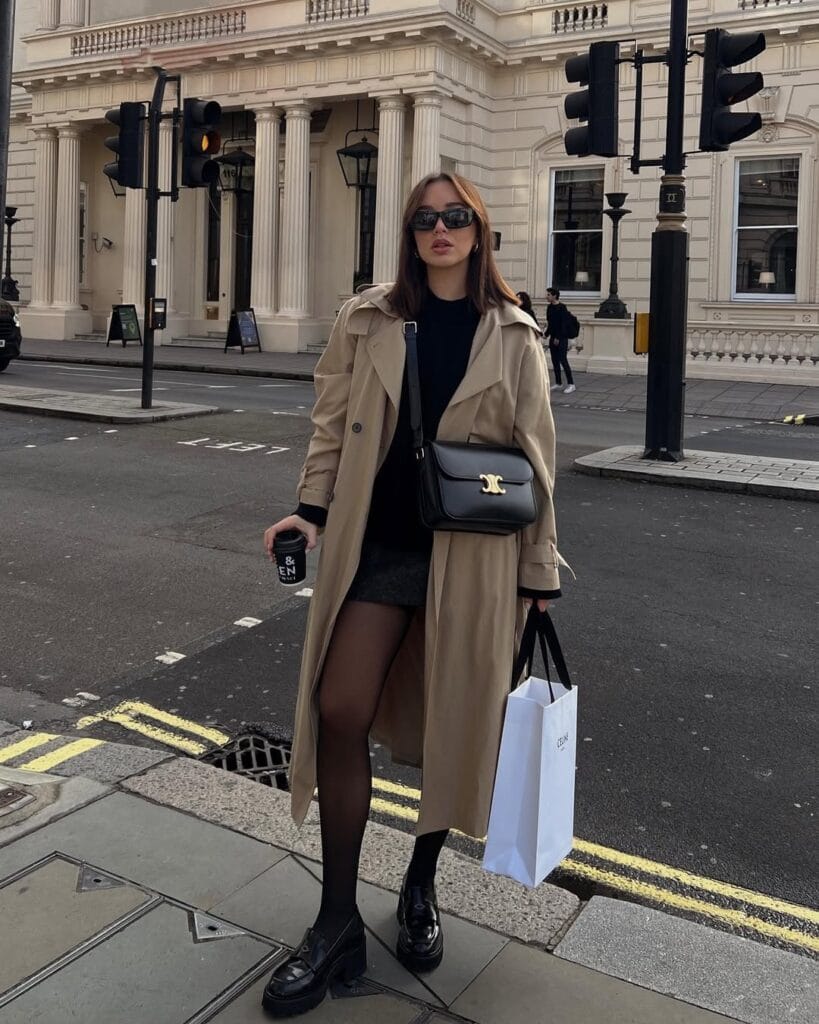 Woman in trench coat, mini skirt, black tights, and loafers crossing a downtown street with shopping bag